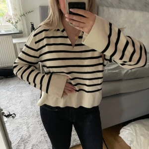 Randig tröja från AWARE XS - Säljer en krämvit och svart randig tröja från AWARE i storlek XS. Tröjan har klassisk krage, v-ringning och långa ärmar med lite vidare avslut. Perfekt för en chill och stilren look. Materialet är mjukt och känns skönt mot huden.