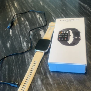Smartwatch Philippe Palmer beige - Snygg smartwatch från Philippe Palmer med fyrkantig urtavla och beige silikonarmband. Klockan har en stor digital display och silverfärgad ram. Laddkabel och originalförpackning medföljer. Perfekt för dig som gillar teknik och vill ha koll på tiden med stil.