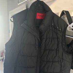 Svart quiltad dunväst från Hugo Boss - Svart dunväst från Hugo Boss med quiltad design och hög krage. Västen har en röd Hugo Boss-logga på insidan och ryggen, samt diskret logotyp på bröstet. Stängs med dragkedja och har en stilren, sportig look. Perfekt för lager-på-lager under kyliga dagar. Den har ett super litet jack på sidan. Nypris 1600