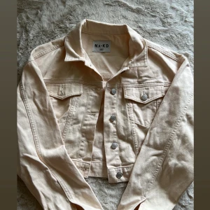 Beige croppad jeansjacka från NA-KD - Snygg croppad jeansjacka från NA-KD i en ljusbeige färg. Jackan har klassisk krage, silverfärgade knappar framtill och två bröstfickor med lock. Perfekt för dig som vill ha en trendig och stilren look.