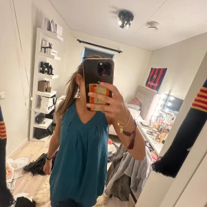 Blå ärmlös linnetopp med v-ringning - Säljer en blå ärmlös linnetopp med v-ringning och veckade detaljer framtill. Toppen har en lös passform och är perfekt för varma dagar. Enkel och stilren design som passar till mycket.