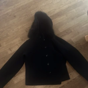 Marinblå kappa med pälskantad huva - Stilren marinblå  kappa med lång ärm och knäppning framtill. Jackan har en stor huva med fluffig pälskant som ger en lyxig känsla. Perfekt för kyliga dagar när du vill hålla dig varm och snygg. Klassisk rak passform och tidlös design. Vi kan distkutera pris 