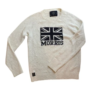 Morris tröja  - Vit stickad tröja från Morris med svart Union Jack-flagga och Morris-logga på bröstet. Tröjan har rund halsringning, långa ärmar och ribbade muddar. Perfekt för dig som gillar klassisk och stilren design.