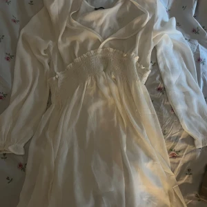 Vit omlottklänning från lager 157 - Säljer en söt vit omlottklänning från lager 157 med långärmade transparenta ärmar och smockad midja med volangdetaljer. Klänningen har v-ringad hals och är gjord i ett lätt, luftigt material som ger en flowy känsla. Perfekt för varma dagar och chill vibes.