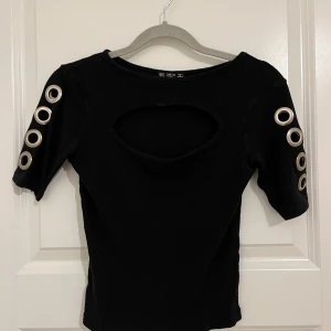 Svart T-shirt med cut-out och metalldetaljer - Cool svart topp från Mistix med korta ärmar och stora silvriga öljetter längs båda ärmarna. Toppen har en snygg cut-out detalj framtill och är ribbad i materialet. Perfekt för dig som vill sticka ut med en edgy stil. Storlek M passar S oxå. 