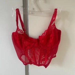 Röd spetskorsett med justerbara band - Säljer en röd korsettopp i spets med transparenta partier och fina broderade detaljer. Toppen har justerbara axelband och hakknäppning bak. Materialet är 95% polyester och 5% elastan vilket gör den stretchig och bekväm. Perfekt för dig som vill ha något extra snyggt i garderoben.