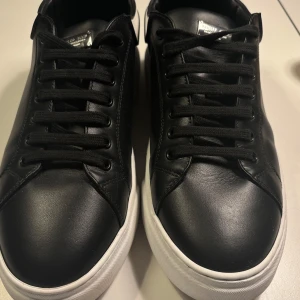 Philipp Plein svarta sneakers i skinn - Svarta sneakers från Philipp Plein i skinn med vit tjock sula och vit hälparti med logga och döskalle. Klassisk rund tå, svarta snören och metallplatta med varumärket på plösen. Snygg kontrast mellan svart och vitt, perfekt för en clean streetstyle.