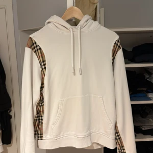 Vit Burberry hoodie med rutiga detaljer - Säljer en vit hoodie från Burberry i 100% bomull med klassiska rutiga paneler på axlar och sidor. Tröjan har huva med snörning, känguruficka och långa ärmar. Perfekt för dig som vill ha en stilren men ändå ikonisk look.