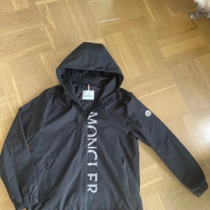 Svart vindjacka från Moncler med huva - Säljer en svart vindjacka från Moncler med huva och dragkedja. Jackan har Moncler-logga på ärmen och stor vit Moncler-tryck längs dragkedjan. Perfekt för blåsiga dagar och enkel att matcha med det mesta.