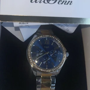 Regal klocka med blå urtavla och stenar - Snygg klocka från Regal med blå urtavla, romerska siffror och tre små urtavlor. Boetten är dekorerad med glittrande stenar runt om och armbandet är i silver och guldtonat metall. Perfekt accessoar för att lyfta din stil.