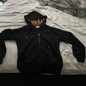 Svart Burberry hoodie med dragkedja pris kan sänkas - Svart hoodie från Burberry med klassiskt rutigt foder i huvan och guldfärgad dragkedja. Tröjan har snörning i huvan, långa ärmar och två fickor framtill. Perfekt för dig som vill ha en stilren och lyxig streetwear-look.