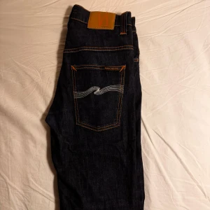 Nudie jeans - Säljer ett par mörkblå jeans från Nudie Jeans, W31 L32 med klassisk orange söm och vågig brodyr på bakfickorna. Modellen är Thin Fin, alltså slim fit/skinny. Perfekt för dig som gillar stilrena och tidlösa jeans. Jeansen är knappt använda, ser ut som nya. Priset kan diskuteras.