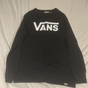 Svart sweatshirt från Vans - Svart sweatshirt från Vans med stort vitt logotryck på bröstet. Klassisk rund halsringning och långa ärmar. Mjuk insida och ribbade muddar vid ärmslut och nederkant. Perfekt för en avslappnad streetstyle-look.