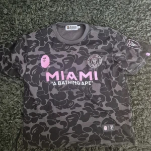 Inter Miami x BAPE camo fotbollströja M - Unik Inter Miami fotbollströja från A Bathing Ape med mörkgrått och svart camouflagemönster. Stora rosa tryck med MIAMI och BAPE 93, samt klubbmärke på bröstet. Klassisk rund hals och korta ärmar. Perfekt för dig som gillar streetwear och fotboll.