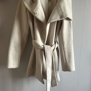 Beige kappa med bälte  - Snygg beige kappa med markerad stor krage och bälte i midjan. Jackan har knappar framtill och är långärmad med en klassisk, stilren look. Perfekt för dig som gillar en clean och elegant stil.