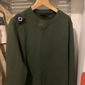 Mörkgrön sweatshirt från MA.STRUM - Snygg mörkgrön sweatshirt från MA.STRUM med klassisk rund hals och lång ärm. Tröjan har en stilren look med diskret logotyp på axeln och är tillverkad i mjuk bomull. Perfekt för en chill och avslappnad stil.