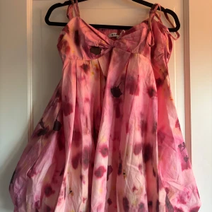 Rosa blommig ballongklänning från NA-KD x Larissa Mills - Superfin kortklänning från NA-KD i återvunnet material. Klänningen har rosa och röda toner med blommigt mönster, knytbara axelband och twistad detalj framtill. Luftig och voluminös passform, perfekt för sommarens varma dagar. Storlek 38 men passar som en 36.