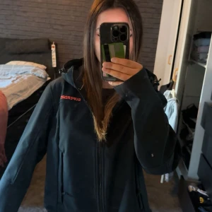 Svart vindjacka från Superdry - Svart vindjacka från Superdry med röd logga på bröstet. Jackan har huva, dragkedja framtill och två sidofickor med dragkedja. Modellen heter Windtrekker Evolution/20 och är perfekt för blåsiga dagar. Snygg och sportig look.