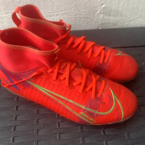 Nike Mercurial röda fotbollsskor - Säljer ett par Nike Mercurial fotbollsskor i en riktigt snygg röd färg med gröna och lila detaljer. Skorna har snörning, högre skaft och mönstrad sula med dobbar för gräsplan. Perfekta för dig som vill sticka ut på planen!