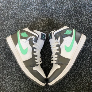 Nike Air Jordan 1 Mid svart/vit mint - Nike Air Jordan 1 Mid sneakers i svart och vitt med mintgröna detaljer. Klassisk high-top siluett med snörning, perforerad tå och ikonisk Swoosh i mint. Ovandel i skinn och syntet, svart foder och vit sula. Perfekta för dig som vill sticka ut med stil.