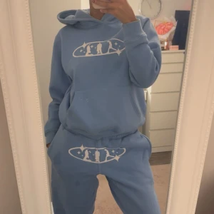 Blå hoodie med tryck och huva - Säljer en ljusblå hoodie med vit grafiskt tryck framtill. Tröjan har huva, magficka och långa ärmar. Mjuk och skön, perfekt för chill dagar. Passformen är loose och materialet känns som bomull eller fleece.