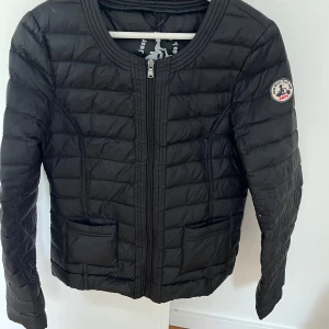 Svart dunjacka från Jott - Säljer en svart, quiltad dunjacka från Jott med rund halsringning och dragkedja framtill. Jackan har två fickor och en snygg patch på ärmen. Perfekt för kyliga dagar och enkel att matcha med allt. Använd vid ett tillfälle.