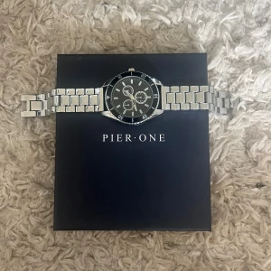 Silverfärgad klocka från Pier One - Stilren klocka från Pier One med svart urtavla och silverfärgat armband i metall. Klockan har flera små urtavlor och tydliga siffror runt boetten. Perfekt accessoar för dig som gillar en klassisk look.