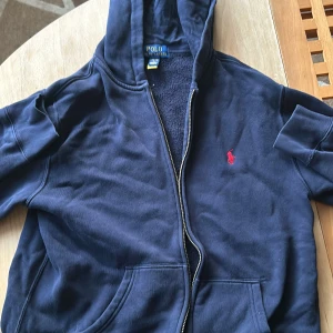 Marinblå hoodie från Polo Ralph Lauren - Snygg marinblå hoodie från Polo Ralph Lauren med dragkedja och huva. Tröjan har det klassiska röda Polo-logot på bröstet, känguruficka och är tillverkad i mjuk bomull. Perfekt för chill dagar eller när du vill ha en clean och stilren look