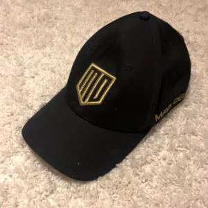 Svart keps från Maya Delorez - Svart keps med böjd skärm och broderad logga i guld framtill. Sidan har texten 'Maya Delorez' i guld. Klassisk form med justerbar rem bak. Perfekt för dig som gillar stilrena accessoarer med en touch av lyx.