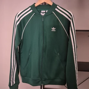 Classic green Adidas zip-up jacket with white stripes. -  Selling a classic green Adidas zip-up jacket with the iconic white stripes along the sleeves and an embroidered logo on the chest. The jacket features a front zipper, two practical zippered pockets, and ribbed cuffs. Perfect for a sporty look or a relaxed everyday style. Completely unworn! :)