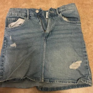 Blå jeanskjol från &Denim - Säljer en blå jeanskjol från &Denim i klassisk modell med slitna detaljer framtill, fem fickor och bälteshällor. Kjolen har dragkedja och knapp framtill och är tillverkad i denim. Perfekt för en avslappnad och cool stil.