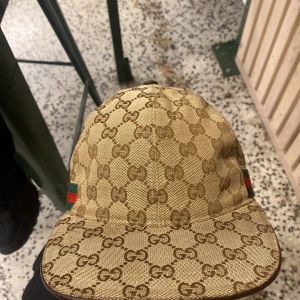 Gucci keps med GG-mönster - Säljer en beige och brun keps från Gucci med klassiskt GG-monogram över hela kepsen. Kepsen har grönt och rött band på sidorna och justerbar rem baktill i brunt skinn. Materialet är tåligt och har en lyxig känsla, perfekt för dig som gillar designeraccessoarer. Självklart äkta.