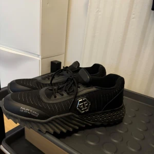 Svarta sneakers från Philipp Plein - Snygga svarta sneakers från Philipp Plein med coola detaljer som silverfärgad logga på sidan och dödskalle på hälen. Skorna har en chunky, mönstrad sula och är gjorda i en mix av textil och skinn. Perfekta för dig som gillar streetstyle och vill sticka ut.