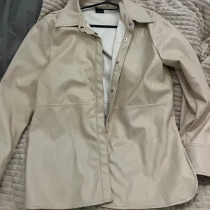 Beige skinnjacka från Gina Tricot - Snygg beige skinnjacka från Gina Tricot i storlek 34. Jackan har klassisk krage, tryckknappar framtill och raka långa ärmar. Materialet är slätt och ger en clean look, perfekt för dig som gillar stilrena vibes. Sparsamt använd🩷