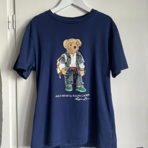 Blå Polo Bear t-shirt från Ralph Lauren 10-12 år - Mörkblå t-shirt från Ralph Lauren med ikoniskt Polo Bear-tryck på bröstet. Klassisk passform med rund hals och korta ärmar. Tillverkad i mjuk bomull, perfekt för en avslappnad och cool stil. Är i mycket bra användt skick, dock så är texten bredvid qrkoden borttvättat.