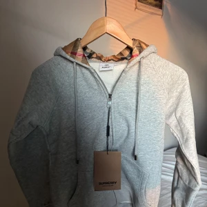 Grå hoodie från Burberry - Säljer en grå hoodie från Burberry med klassiskt rutigt foder i huvan. Tröjan har dragkedja framtill, snörning vid huvan och två fickor. Perfekt för dig som gillar stilrena och exklusiva plagg med en twist.