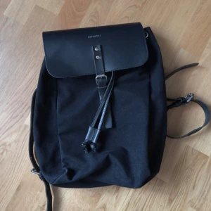 Svart stilren ryggsäck med spänne Sandqvist - Snygg svart ryggsäck med minimalistisk design och lock i skinn. Väskan har ett klassiskt spänne framtill och praktiskt handtag upptill. Perfekt för dig som gillar enkel och stilren look.