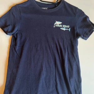 Mörkblå Shark Squad t-shirt Pepperts - Mörkblå t-shirt från Pepperts med coolt Shark Squad-tryck och två hajar på bröstet. Klassisk rund hals och korta ärmar. Skön och mjuk i bomull, perfekt för dig som gillar havet och vill sticka ut lite extra.