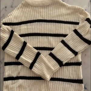 Randig stickad tröja från 157 - Vit stickad tröja med svarta horisontella ränder från 157. Tröjan har en högre ribbad hals, långärmade ärmar och en loose fit. Perfekt för lager-på-lager och chill höst- eller vinterstil.