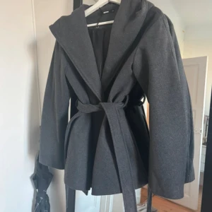 Mörkgrå kappa med huva och bälte - Snygg mörkgrå kappa från Bik Bok med stor huva och knytskärp i midjan. Perfekt för dig som vill ha en stilren och mysig look. Nypris 800kr 