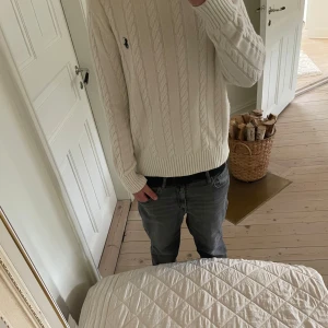 Vit kabelstickad tröja från Polo Ralph Lauren - Säljer en vit kabelstickad tröja från Polo Ralph Lauren med klassisk blå broderad logga på bröstet. Tröjan har rund hals, ribbade muddar och lång ärm. Perfekt för dig som gillar stilrena och tidlösa plagg med snygg passform.
