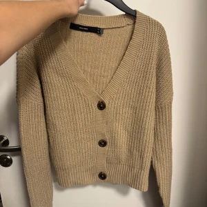 Beige stickad kofta från Vero Moda - Beige stickad kofta från Vero Moda med grov ribbstickning, djup v-ringning och tre stora knappar framtill. Långärmad och lite boxig passform, perfekt att slänga över en t-shirt eller linne. Enkel och stilren design som funkar till allt.