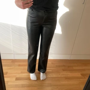 Svarta ”skinnbyxor” från NA-KD, stl 38 - Säljer ett par svarta ”skinnbyxor” från NA-KD i storlek 38. Byxorna har rak passform, hög midja och stängs med dragkedja och knapp. Snyggt material med en stilren look, perfekta för att lyfta din outfit.