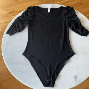 Svart ribbad body med puffärm - Svart ribbad body med rund halsringning och snygga puffärmar i trekvarts längd. Tight passform som framhäver siluetten och ger en trendig look. Perfekt att styla med jeans eller kjol för en cool outfit.