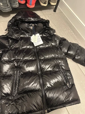 Svart Moncler pufferjacka med huva - Säljer en svart, glansig pufferjacka från Moncler med huva och dragkedja framtill. Jackan har två snedställda fickor med dragkedja och Moncler-logga på ärmen. Perfekt för kalla dagar och riktigt snygg streetstil. Passar runt 180-185