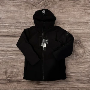 Svart dunjacka från Sail Racing helt ny - Svart dunjacka från Sail Racing med huva och hel dragkedja framtill. Jackan är vindtät, vattenavvisande och har RDS-certifierat dun. Snygg, stilren design med logga på bröstet och huvan. Perfekt för kalla dagar och har en modern look.