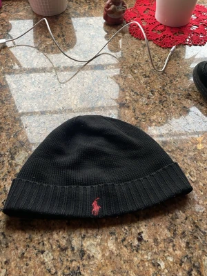Svart mössa från Polo Ralph Lauren - Svart ribbstickad mössa från Polo Ralph Lauren med klassisk röd broderad logga framtill. Mössan är tillverkad i mjuk bomull och har en enkel, stilren design som passar perfekt till kalla dagar.