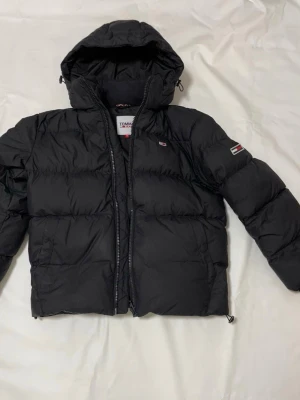 Svart pufferjacka från Tommy Jeans - Svart pufferjacka från Tommy Jeans med huva och dragkedja framtill. Jackan har broderad logga på bröstet och är quiltad för extra värme. Perfekt för kalla dagar och har en clean, modern look som funkar till streetwear.