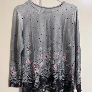 Grå långärmad blommig tröja - Säljer en grå långärmad tröja med blommigt mönster i rosa, vinrött och beige. Tröjan har rund hals och ett detaljerat tryck med små blad och blommor som går från mitten och nedåt. Materialet känns mjukt och lätt, perfekt för dig som gillar unika prints.