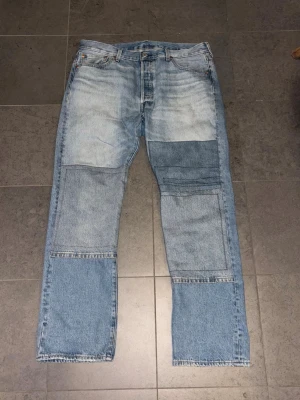 Patchade Levi's 501 jeans ljusblå - Unika Levi's 501 jeans i ljusblå tvätt med flera stora patchade detaljer på framsidan av benen. Klassisk femficksmodell med raka ben och knappgylf. Jeansen är tillverkade i slitstark bomull och har den ikoniska läderlappen bak i midjan.